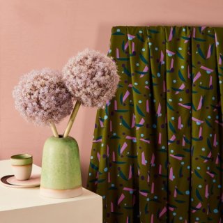 Die Ansicht zeigt den Viscosestoff Java ivy green weich fliessend über eine Stange gehaengt, welche an der Wand befestigt ist. Es ist ein Stoffdruck mit blauen und lila Punkten, rosa und pertrolfarbenen Strichen auf avocadogruenen Untergrund.