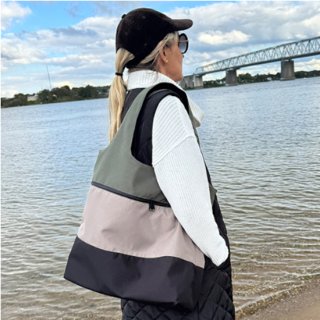 Das Bild zeigt eine Frau mit Blick auf eine Bruecke, die ueber einen Fluss fuehrt. Sie traegt dabei ein dreifarbige Tasche ueber der rechten Schulter. Die Dreifarbigkeit entsteht durch drei verschiedenfarbige zusammengenaehte Stoffe. Der obere und mittlere Stoff sind durch einen Reißverschluss geteilt. Die Taschenfarben sind mint, beige und dunkelblau.