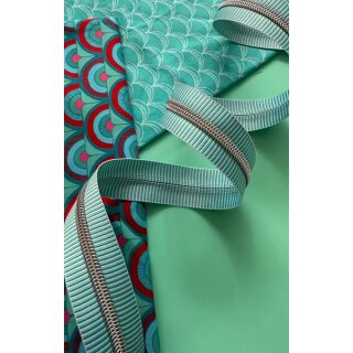 Das Bild zeigt den Endlos-Reissverschluss tuerkis weisse Streifen mit nickelfarbenen Zaehnen. Dekoriert ist er vor einer Vielzahl an bunten Stoffen.