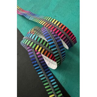 Das Bild zeigt den Endlos-Reissverschluss regenbogen schwarze Blockstreifen mit nickelfarbenen Zaehnen. Dekoriert ist er vor einer Vielzahl an bunten Stoffen.